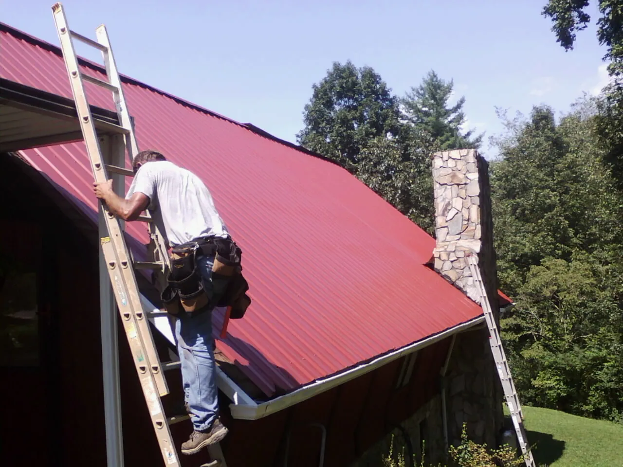 Roofer on a ladder beside a red metal roof and stone chimney — Soffit & Fascia Repair by Roofing Brothers in Oceanport